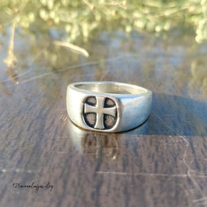 Pu&ograve; includere: Anello d'argento con design a croce. L'anello ha una fascia larga e una faccia ovale rialzata con una croce al centro. L'anello &egrave; realizzato in un materiale metallico lucido ed &egrave; probabilmente un gioiello.