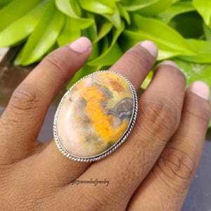 May include: An oval-shaped ring featuring a large, polished bumblebee jasper stone with swirling patterns of yellow, orange, gray, and white. The stone is set in a silver bezel with a twisted rope-style border. The text 925moonleafjewelry is visible.