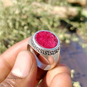 Può includere: Un anello d'argento con una grande gemma rossa ovale. L'anello ha un bordo decorativo con un motivo geometrico ripetuto. L'anello è tenuto in mano su uno sfondo verde sfocato. Il testo "125moonleafjewelry" è visibile.