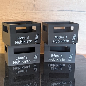 Könnte beinhalten: Vier schwarze Metall-Aufbewahrungsboxen, die auf einer reflektierenden Oberfläche gestapelt sind. Jede Box hat einen Namen und eine Flaschenillustration in Weiß. Der Text auf den Boxen lautet: "Hero's Hubikiste", "Micha's Hubikiste", "Ihno's Hubikiste" und "Efen's Hubikiste".