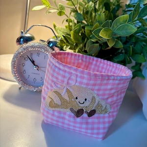 May include: A pink gingham fabric basket with an embroidered croissant design. A silver alarm clock with a crystal-studded bezel is in the background, along with green leafy plants. The basket is a small, decorative storage item.