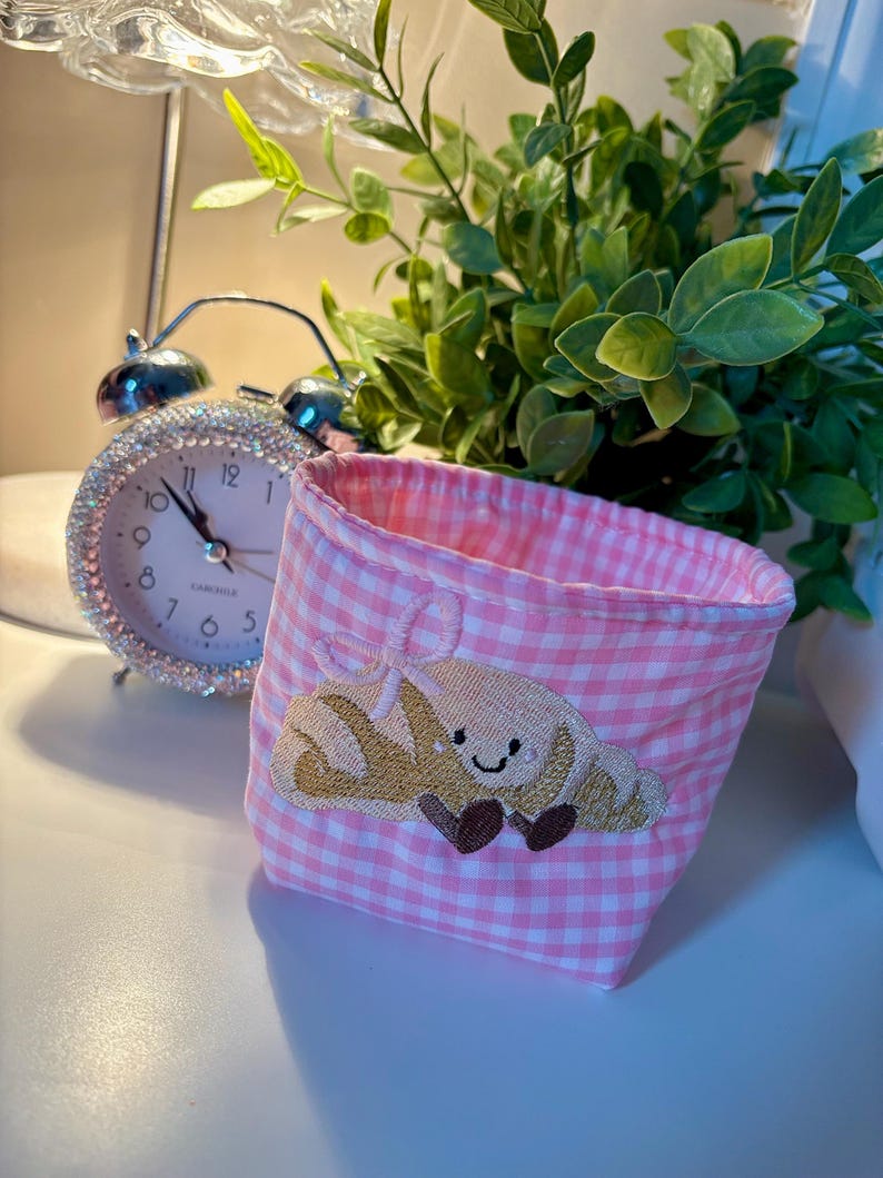 May include: A pink gingham fabric basket with an embroidered croissant design. A silver alarm clock with a crystal-studded bezel is in the background, along with green leafy plants. The basket is a small, decorative storage item.