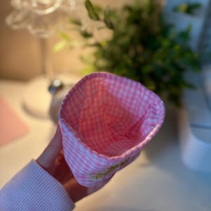 May include: A pink and white gingham fabric hat with a gold embroidered detail. The interior of the hat is also pink and white gingham. The hat is held by a person wearing a pink sweater.