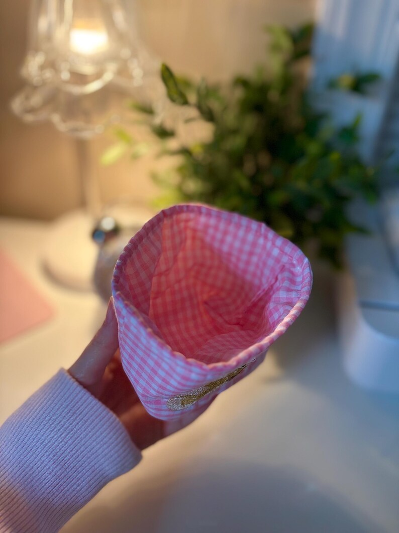 May include: A pink and white gingham fabric hat with a gold embroidered detail. The interior of the hat is also pink and white gingham. The hat is held by a person wearing a pink sweater.