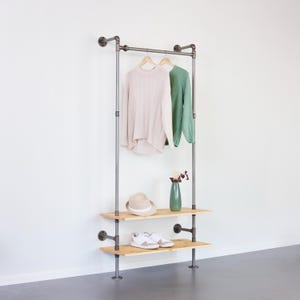 Pode incluir: Cabideiro de estilo industrial feito de tubos de metal com duas prateleiras de madeira. Um suéter bege e um suéter verde penduram na barra superior. Um chapéu, um vaso verde e tênis brancos estão nas prateleiras. O cabideiro está contra uma parede branca.