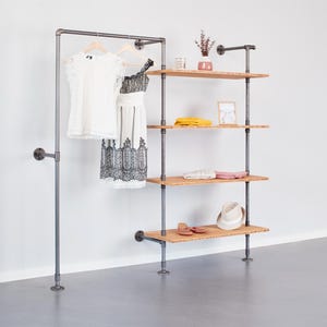Könnte beinhalten: Ein Kleiderständer und ein Regal aus grauem Metall und Holz. Der Ständer hat eine Kleiderstange mit zwei weißen Kleidern. Die Regale zeigen gefaltete Kleidung, einen Hut, Schuhe und dekorative Gegenstände. Die Einheit steht vor einer weißen Wand.