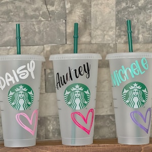 May include: Three clear plastic cold cups with lids and green straws. Each cup has a name in a different font: "Daisy", "Audrey", and "Michele". Each cup has a heart graphic in pink or purple. The Starbucks logo is visible.