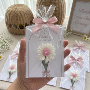 May include: A baby shower favor featuring a white and pink daisy-shaped item on a card, wrapped in clear cellophane and tied with a pink ribbon. The card reads "Rosie's Baby Shower". Several favors are visible.
