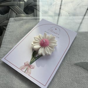 May include: A white daisy-shaped candle with a pink centre sits on a card. The card features the text "Rosie's Baby Shower" and a watercolour illustration of a daisy stem and bow. The background is grey fabric.
