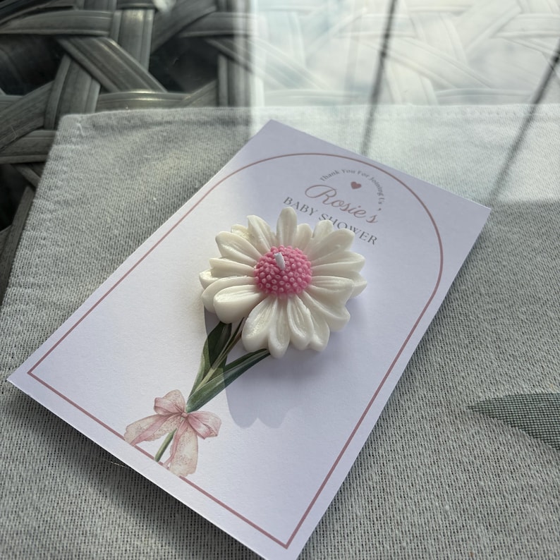 May include: A white daisy-shaped candle with a pink centre sits on a card. The card features the text "Rosie's Baby Shower" and a watercolour illustration of a daisy stem and bow. The background is grey fabric.