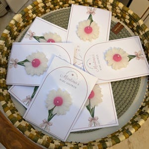 May include: Baby shower invitations with a white daisy candle and pink centre. The cards are white and feature the text "Martina's Baby Shower" and a decorative bow. The invitations are arranged in a woven basket.