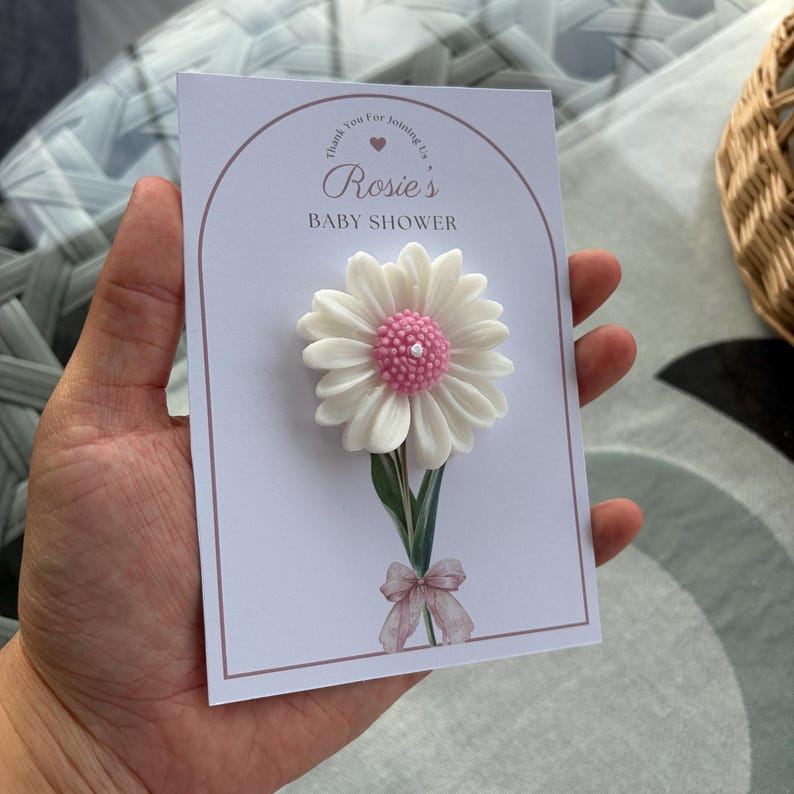 May include: A baby shower card with a large white and pink daisy. The card reads "Rosie's BABY SHOWER" in script, with a decorative bow at the base of the flower. The card is held in a hand.