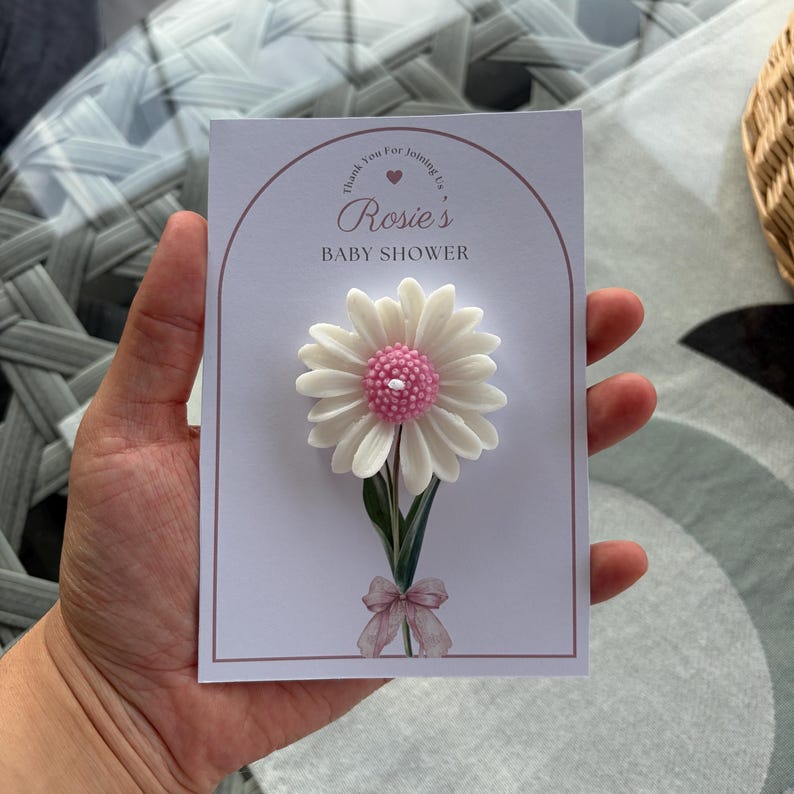 May include: A baby shower invitation card with a 3D daisy design. The daisy has white petals and a pink centre. The card reads "Rosie's BABY SHOWER" with a decorative arch and a small bow at the base of the flower.