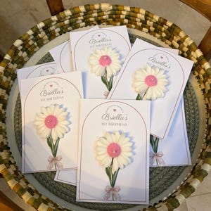 May include: Several white cards, each featuring a daisy candle. The cards are printed with the text "Briella's 1st Birthday" in a pink arch. The daisy candles have white petals and a pink centre, with a green stem and a pink bow.