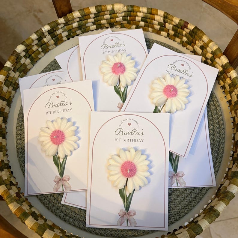 May include: Several white cards, each featuring a daisy candle. The cards are printed with the text "Briella's 1st Birthday" in a pink arch. The daisy candles have white petals and a pink centre, with a green stem and a pink bow.