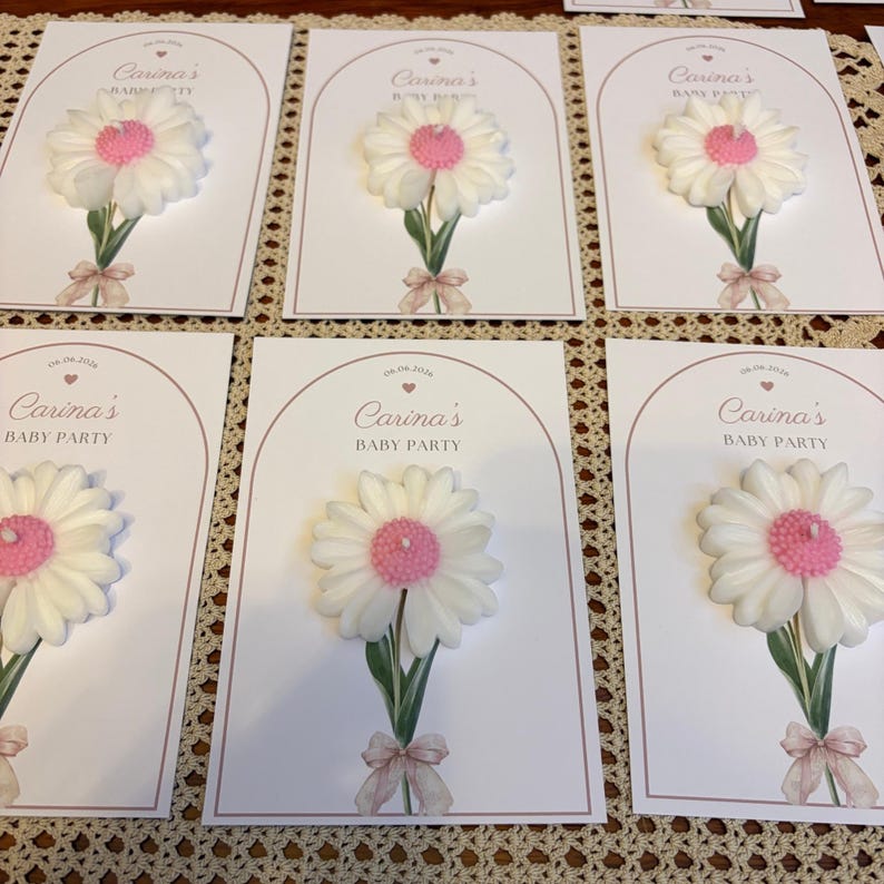 May include: Baby shower invitations with a white daisy and pink centre, green stem, and a pink bow. The cards read "Carina's BABY PARTY" and the date "06.06.2024". The cards are arranged on a patterned surface.