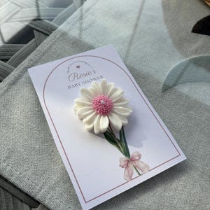 May include: A white daisy-shaped candle with a pink centre sits on a card. The card reads "Rosie's BABY SHOWER" in pink script. A watercolour bow and greenery accent the card's design. The card is on a grey surface.