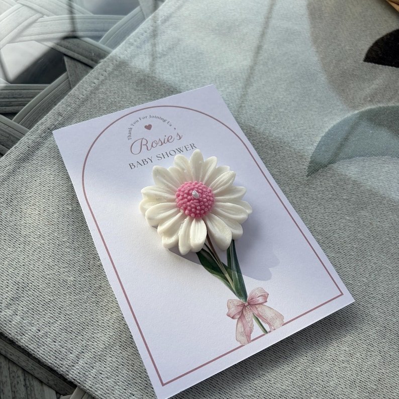 May include: A white daisy-shaped candle with a pink centre sits on a card. The card reads "Rosie's BABY SHOWER" in pink script. A watercolour bow and greenery accent the card's design. The card is on a grey surface.