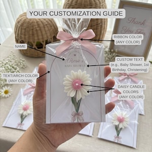 May include: A daisy candle, with white petals and a pink centre, packaged in cellophane and tied with a pink ribbon. The card reads "Rosie's Baby Shower" and offers customisation options for text, colours, and ribbon.
