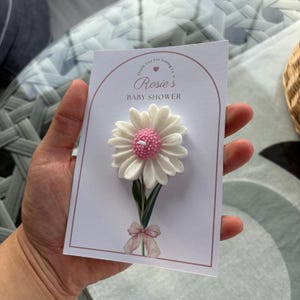May include: A baby shower invitation card with a white daisy candle, pink centre, and green stem. The card reads "Rosie's BABY SHOWER" in a decorative font, with a pink bow at the base of the flower.
