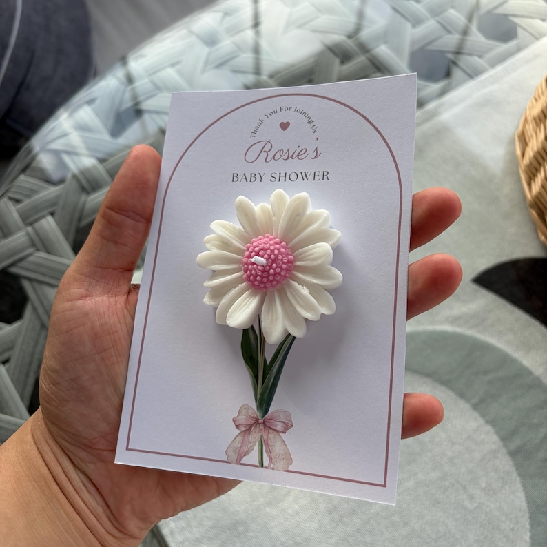 May include: A baby shower invitation card with a white daisy candle, pink centre, and green stem. The card reads "Rosie's BABY SHOWER" in a decorative font, with a pink bow at the base of the flower.