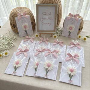 May include: Baby shower favours with daisy decorations, individually wrapped and tied with pink ribbons. A sign reads "Thank You For Coming". A woven basket and daisies are also visible, creating a celebratory atmosphere.