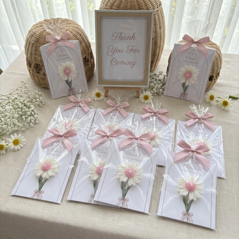 May include: Baby shower favours with daisy decorations, individually wrapped and tied with pink ribbons. A sign reads "Thank You For Coming". A woven basket and daisies are also visible, creating a celebratory atmosphere.