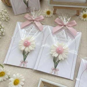 May include: Baby shower favours featuring daisy-shaped candles with pink centres and green stems, attached to cards with the text "Rosie's BABY SHOWER". Each favour is wrapped in clear cellophane and tied with a pink ribbon.