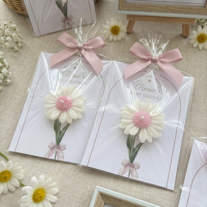 May include: Baby shower favours featuring daisy-shaped candles with pink centres and green stems, attached to cards with the text "Rosie's BABY SHOWER". Each favour is wrapped in clear cellophane and tied with a pink ribbon.