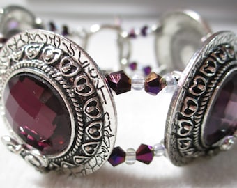 May include: A silver bracelet with two large round charms featuring a purple gemstone surrounded by a heart design. The bracelet is accented with small purple and clear glass beads.