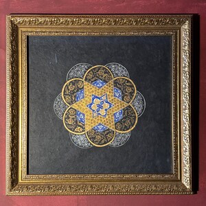 Puede incluir: Una obra de arte enmarcada con un diseño simétrico en oro, azul y blanco sobre un fondo negro. La obra de arte está en un marco dorado y detallado, creando una estética clásica. El diseño tiene forma de estrella con patrones florales y geométricos.