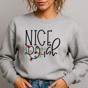 May include: A grey sweatshirt featuring the text "NICE Wish" in black with a festive string of lights. The crew neck top is worn with blue jeans. The image is of a person.
