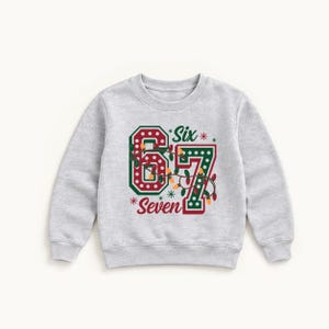 May include: A light grey crewneck sweatshirt featuring a large graphic of the numbers 6 and 7 in red and green with white polka dots. The design includes a string of colourful Christmas lights draped over the numbers and the text Six Seven.