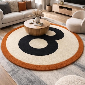 以下が含まれることがあります： クリーム色の背景に大きな黒い数字の8がデザインされた丸いラグ。オレンジ色の縁取りがあり、リビングルームの模様入りのグレーのラグの上に配置されています。直径は約180cmです。