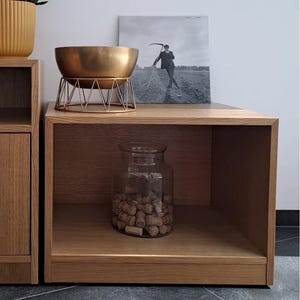 Pode incluir: Uma tigela de metal dourado em um suporte geométrico, uma foto em preto e branco emoldurada, um frasco de vidro cheio de rolhas e uma planta em vaso. Os itens são exibidos em um armário de madeira.