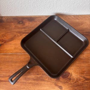 May include: A dark brown cast iron divided griddle pan with a handle. The pan is divided into three sections. The pan is on a wooden surface.