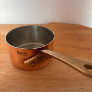 Può includere: Una casseruola color rame con manico in ottone. L'interno è in acciaio inossidabile. Il manico è fissato con rivetti. La scritta "Mauviel Made in France" è sul lato. Un articolo per la cucina.