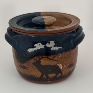 May include: A ceramic jar with a lid, featuring a deer silhouette against a brown and blue landscape. The jar has a dark blue rim and handles, with a two-tone lid in brown and blue. The jar is approximately 10 cm tall.