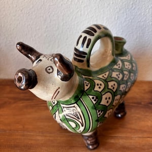 May include: A ceramic animal-shaped vessel featuring a cream-colored head with dark brown ears and snout. The body is decorated with a green and white scale pattern, dark brown legs, and a curved handle on the back with black horizontal stripes.