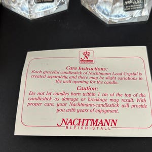 May include: A close-up of a white card with red text and a logo. The card provides care instructions for Nachtmann Lead Crystal candlesticks, including a caution about candle usage. Two crystal candle holders are visible in the background.