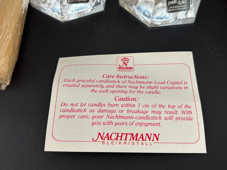 May include: A close-up of a white card with red text and a logo. The card provides care instructions for Nachtmann Lead Crystal candlesticks, including a caution about candle usage. Two crystal candle holders are visible in the background.