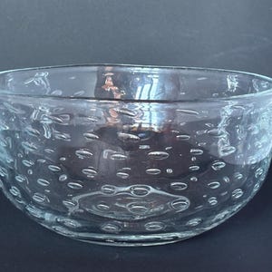 Könnte beinhalten: Eine klare Glasschale mit strukturierter Oberfläche. Die Schale hat eine breite, abgerundete Form und ist mit zahlreichen kleinen, blasenartigen Vertiefungen verziert. Das Glas scheint dick und robust zu sein, geeignet zum Servieren oder Ausstellen.