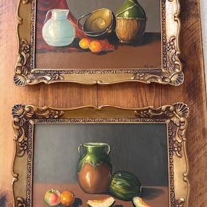 Può includere: Due dipinti di nature morte incorniciati. Il dipinto in alto presenta una brocca azzurra, un secchio dorato, arance e una bottiglia verde. Il dipinto in basso mostra un vaso verde e marrone, un melone e frutta. Entrambi i dipinti hanno cornici dorate ornate.