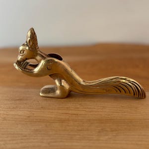 Könnte beinhalten: Ein Nussknacker aus Messing in Form eines Eichhörnchens. Das Eichhörnchen sitzt und hält seine Pfoten hoch. Der Schwanz ist lang und gebogen. Der Nussknacker ist aus Messing und hat eine glänzende Oberfläche.