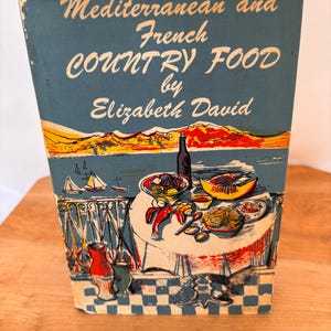 Può includere: Libro di cucina vintage intitolato "Mediterranean and French Country Food by Elizabeth David". La copertina presenta un'illustrazione costiera con barche a vela, un tavolo imbandito con cibo e una bottiglia di vino. Formato verticale.