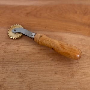 Pode incluir: Uma roda de pastelaria com cabo de madeira e uma lâmina de corte de latão em forma de estrela. O cabo é castanho claro e a lâmina é fixada a uma peça de metal prateado. A ferramenta foi concebida para cortar massa.