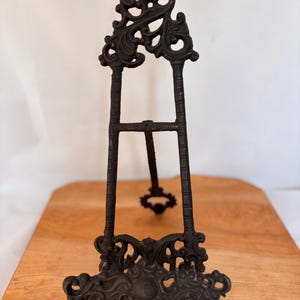 Puede incluir: Un caballete de metal negro con adornos ornamentados y una parte superior decorativa. El caballete tiene un diseño de tres patas y es adecuado para exhibir obras de arte o fotografías. El caballete mide aproximadamente 30 cm de alto.