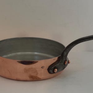 Può includere: Una padella color rame con un manico scuro e curvo. L'interno della padella è argentato. Il manico è fissato con rivetti. Questa padella è progettata per cucinare e misura circa 20 cm di diametro.