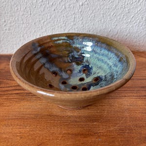 Peut inclure: Un bol à baies en céramique ronde avec une glaçure brillante aux tons mélangés de brun terreux, de bleu marine profond et de bleu sarcelle doux. La base intérieure du bol comporte plusieurs petits trous de drainage régulièrement espacés.