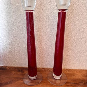 May include: A pair of tall, slender candlesticks made of deep red glass with a subtle speckled texture. Each candlestick features a clear glass circular base and a clear glass cup at the top designed to hold a candle, standing upright.